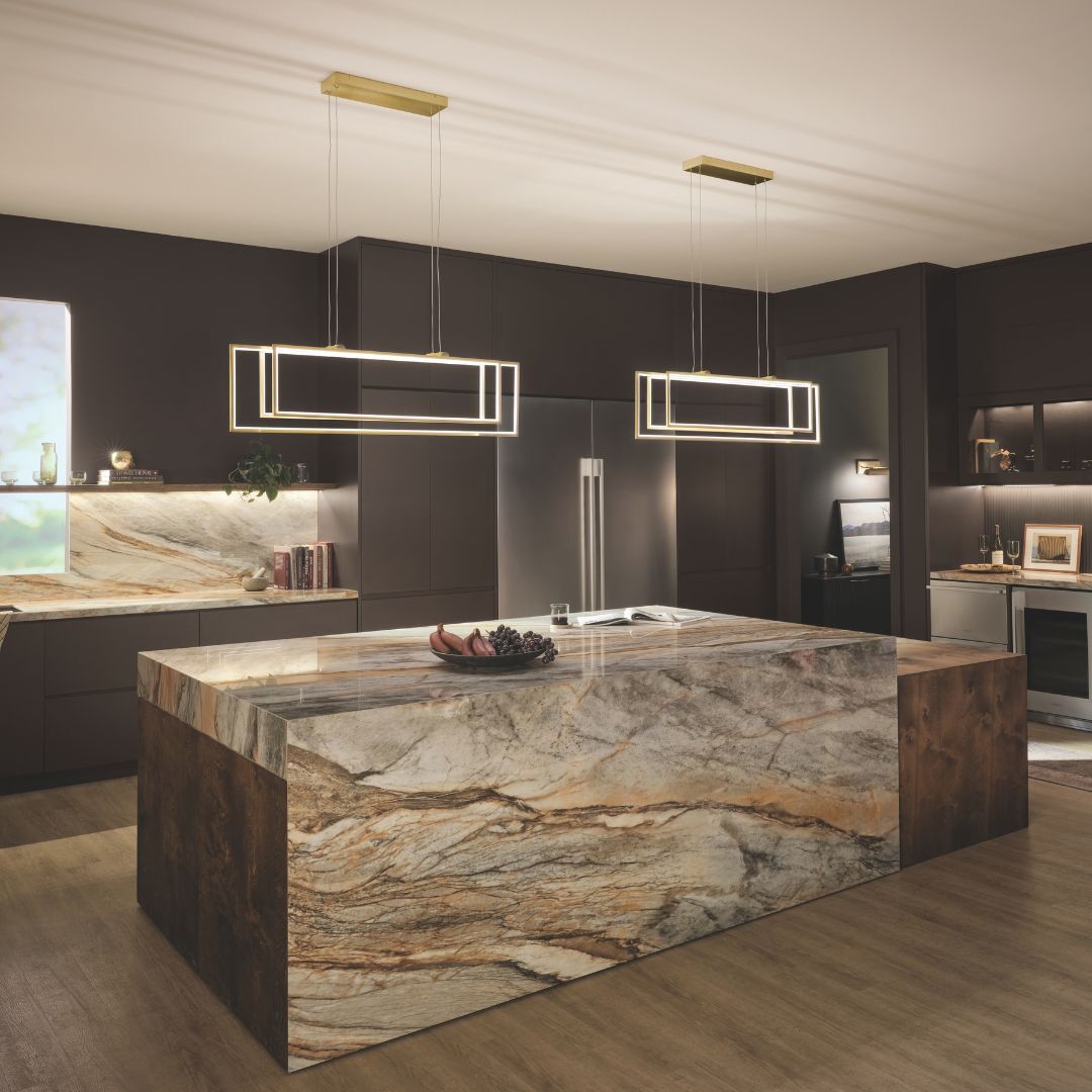 Two modern, rectangular LED pendant lights with a gold-toned finish hang horizontally over a kitchen island. The fixtures feature a minimalist, layered geometric frame design that emits a warm, continuous glow.