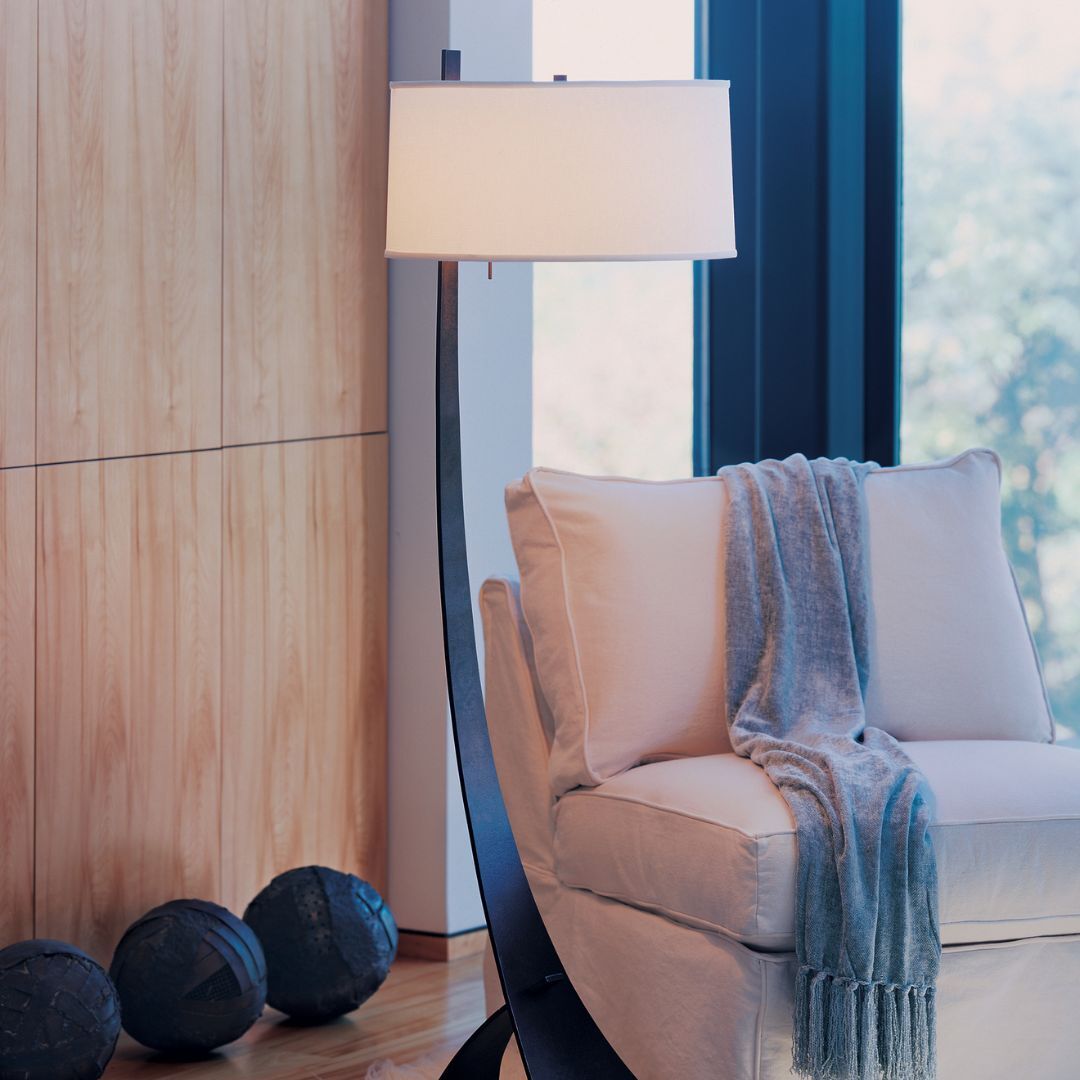 A contemporary floor lamp with a slender, curved black metal body. It is topped with a large, cylindrical white fabric drum shade that provides soft, diffused illumination over a seating area.
