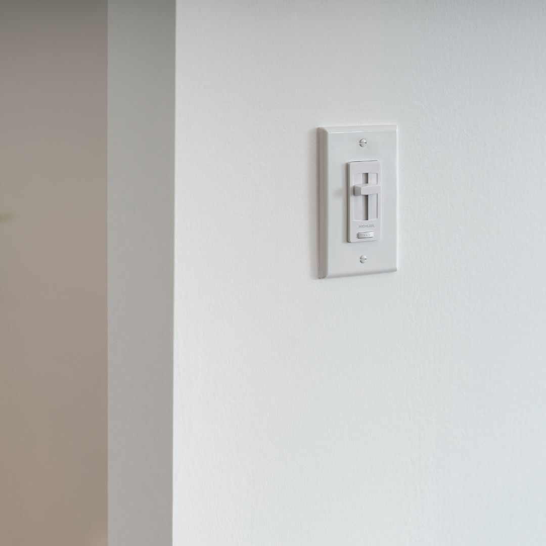 A white, single-pole dimmer switch mounted on a plain wall. The switch features a vertical sliding toggle for brightness control and a small on/off button at the bottom.